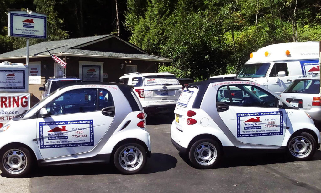 Two white Smart cars and multiple other vehicles parked side by side, each featuring the We Honey Do logo.